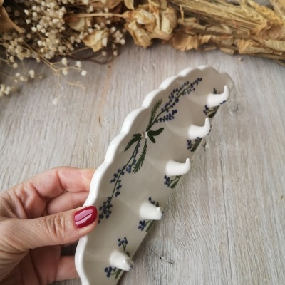 Gancho cerâmico branco decorado com flores azuis e verdes, com 5 ganchos, segurado por mão com unhas vermelhas sobre superfície de madeira.
