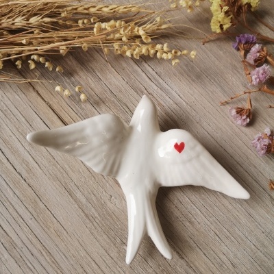Figura cerâmica branca de pássaro com coração vermelho sobre madeira e flores secas
