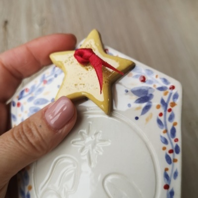 Placa decorativa de cerâmica branca com relevo natalício e estrela amarela com fita vermelha.
