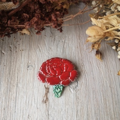 Broche de flor vermelha com haste verde em superfície de madeira clara com flores secas ao fundo