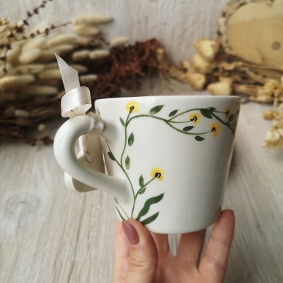 Cana de cerâmica branca com padrão de flores amarelas e folhas verdes e fita branca na asa.