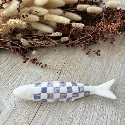 Peixe decorativo em cerâmica com padrão de quadrados azul, junto a ramos secos sobre madeira cinza.