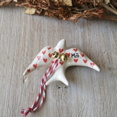 Pendente cerâmico em forma de andorinha branca com corações vermelhos e a palavra 'Mãe', com sinos dourados e corda colorida.