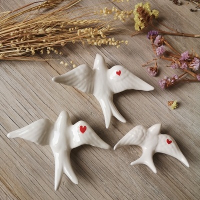 Três figuras cerâmicas em forma de pássaros brancos com corações vermelhos sobre madeira e flores secas