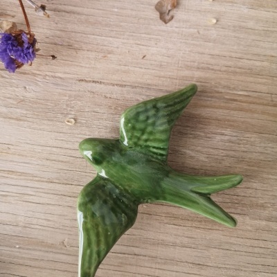 Pássaro verde decorativo em cerâmica sobre madeira clara com flores secas roxas