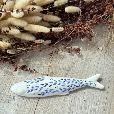 Peixe decorativo em cerâmica branca com padrão azul sobre madeira com flores secas ao fundo