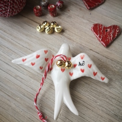 Anjo decorativo branco com corações vermelhos, campainhas douradas e cordão vermelho e branco em superfície de madeira.