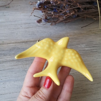 Peça decorativa amarela em forma de pássaro segurada por uma mão com unhas vermelhas sobre mesa de madeira