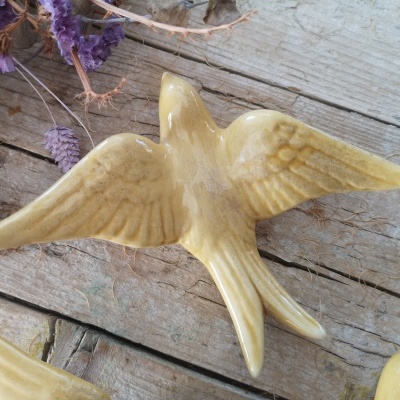 Figura cerâmica de pássaro amarelo com asas abertas sobre madeira rústica e flores secas