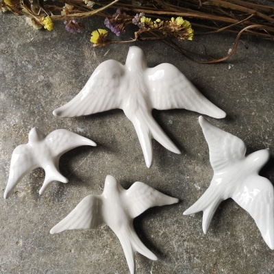 Quatro pássaros decorativos em cerâmica branca num fundo de pedra com flores secas ao lado