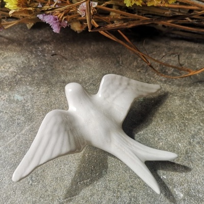 Pássaro decorativo branco em cerâmica com asas abertas sobre superfície cinzenta e flores secas