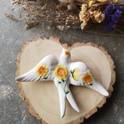 Pássaro decorativo branco em cerâmica pintado com girassóis sobre base de madeira e flores secas ao lado.