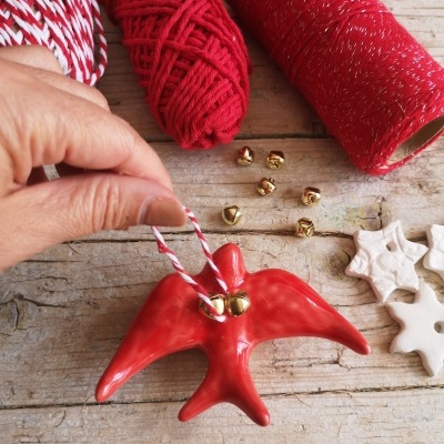Enfeite vermelho em forma de anjo com sinos dourados e novelos de fio vermelho e branco sobre mesa de madeira