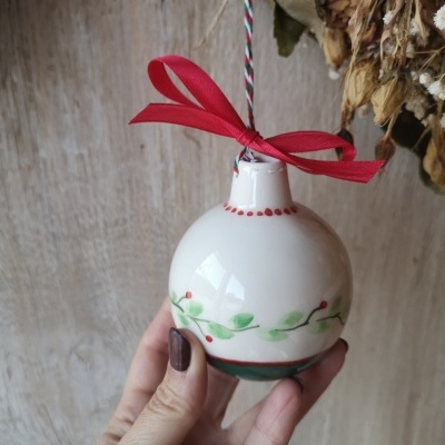 Bola de cerâmica branca decorada com ramos verdes e bagas vermelhas, com fita vermelha e cordão para pendurar.