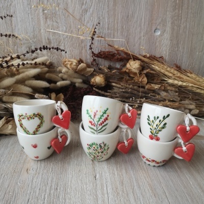 Seis chávenas de porcelana com desenhos coloridos e corações vermelhos nas pegas, sobre madeira clara e flores secas ao fundo.