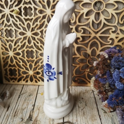 Estatueta branca de figura religiosa em cerâmica com decoração azul, sobre fundo decorativo de madeira e flores secas.