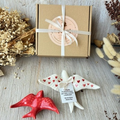 Caixa castanha com laço branco e duas figuras de anjos de cerâmica, uma branca com corações vermelhos e outra vermelha, decoradas com ramos secos