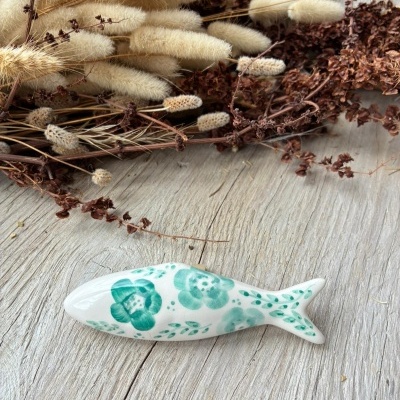 Figura de peixe em cerâmica branca com flores verdes junto de flores secas sobre mesa de madeira clara