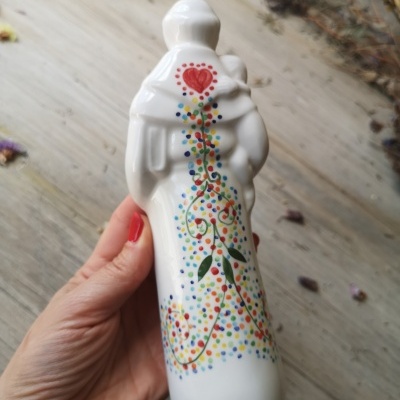 Figura cerâmica branca decorada com padrão colorido e coração vermelho, segurada à mão com unhas vermelhas.