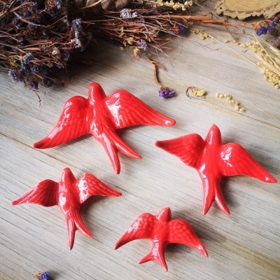Quatro estatuetas vermelhas de pássaros em cerâmica sobre madeira clara com flores secas ao redor