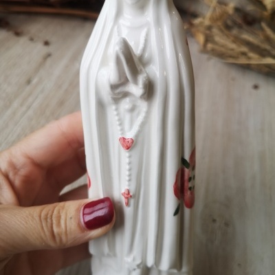 Estatueta religiosa branca em porcelana com terço vermelho e detalhes florais, segurada por mão com unhas vermelhas