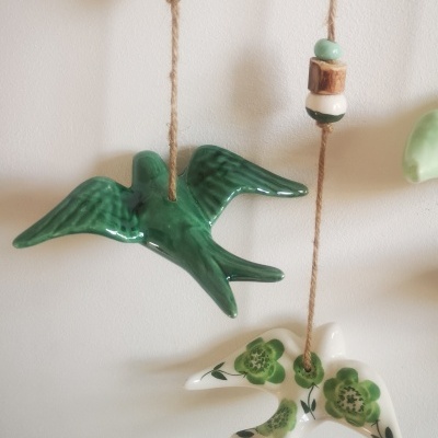 Dois pássaros decorativos de cerâmica suspensos em cordas com contas, um verde e outro branco com flores verdes