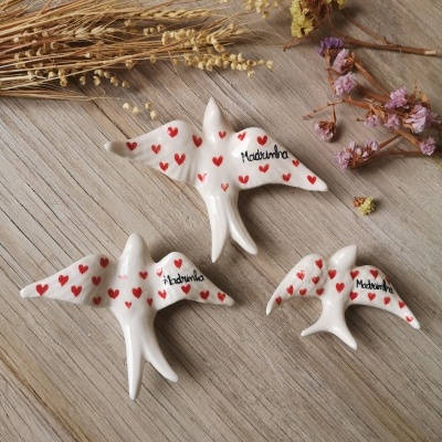 Três pássaros de cerâmica branca com corações vermelhos e texto 'Madeirinha' sobre madeira clara com ramos secos