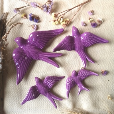 Quatro pássaros decorativos em cerâmica roxa sobre tecido bege com flores secas