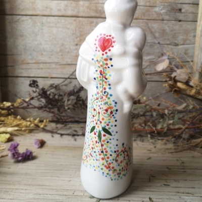 Escultura branca de cerâmica com figura e criança, decorada com pontos coloridos, sobre superfície de madeira rústica