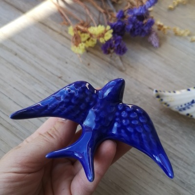 Peça de cerâmica azul em forma de pássaro sobre madeira clara com flores secas ao fundo