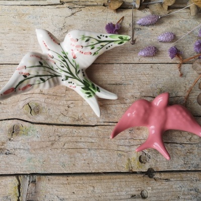 Dois pássaros decorativos em cerâmica branca com padrões florais e rosa sólido sobre madeira rústica.
