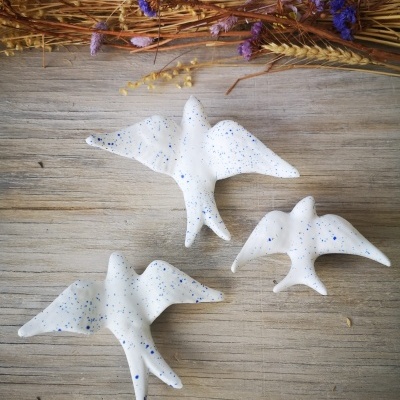 Três pássaros decorativos brancos com pintas azuis sobre madeira clara com flores secas ao fundo