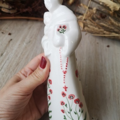 Estatueta de cerâmica branca com flores vermelhas e folhas verdes, detalhe de rosário vermelho, em mão feminina com unhas vermelhas