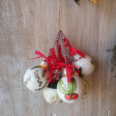 Conjunto de bolas de Natal em cerâmica branca com desenhos de folhas e bagas, com fitas vermelhas, penduradas contra um fundo de madeira clara.