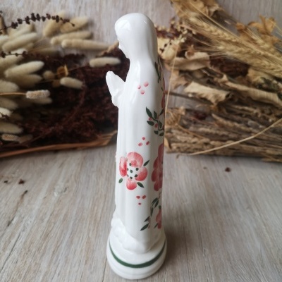 Estátua religiosa branca de porcelana com flores vermelhas e verdes, em uma mesa de madeira clara com flores secas ao fundo.