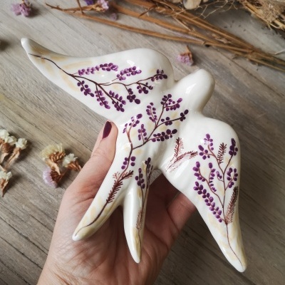 Escultura cerâmica pássaro branco com pintura roxa, mão segurando, flores secas fundo