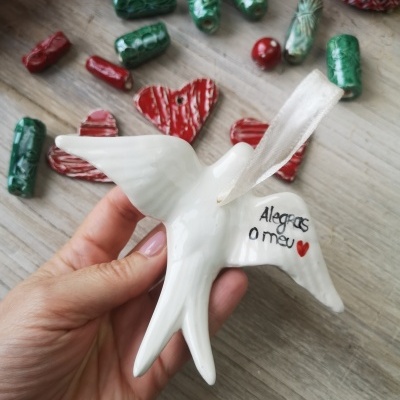 Pássaro decorativo branco em cerâmica com fita para pendurar e mensagem escrita, com outras peças decorativas vermelhas e verdes ao fundo.