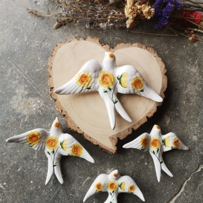 Figuras decorativas de aves em cerâmica branca com flores amarelas, sobre base de madeira e flores secas.