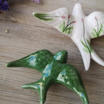 Duas figuras de pássaros em cerâmica, uma verde e outra branca com desenhos florais, sobre madeira