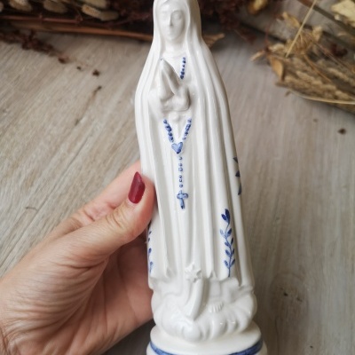 Figura religiosa branca com detalhes azuis de terço e rosas, mão segurando, fundo de madeira e ramos secos