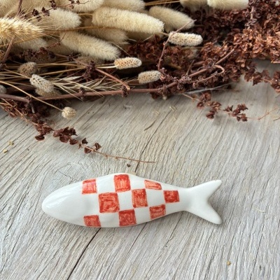 Peixe decorativo em cerâmica branca com padrão xadrez vermelho sobre madeira clara com flores secas.
