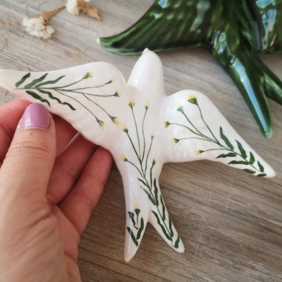 Figura de pássaro em cerâmica branca com pintura verde e amarela sendo segurada à mão sobre superfície de madeira