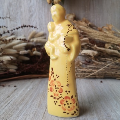 Estatueta de cerâmica amarela com decorações florais e terço, em fundo de elementos naturais secos