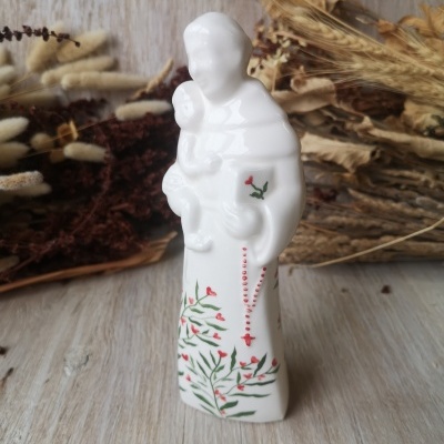 Estátua religiosa branca de porcelana com decoração floral e rosário vermelho, em fundo de plantas secas sobre madeira clara