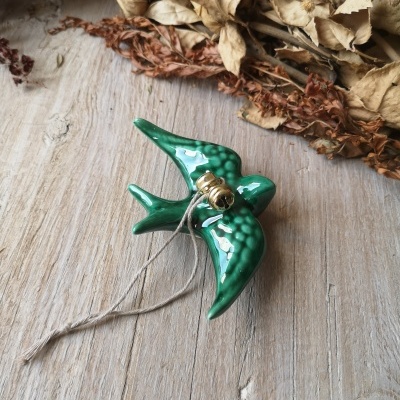 Figura de pássaro em cerâmica verde com sino dourado e fio, sobre madeira clara com folhas secas