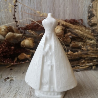 Estatueta religiosa branca em cerâmica com manto e coroa sobre madeira clara e plantas secas