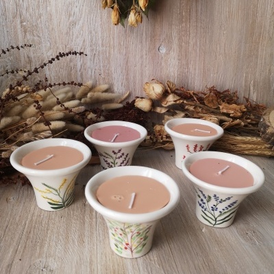 Cinco velas em recipientes de cerâmica branca com desenhos florais em madeira com flores secas ao fundo
