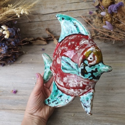 Peixe decorativo em cerâmica pintada à mão em tons vermelhos e verdes sobre madeira clara
