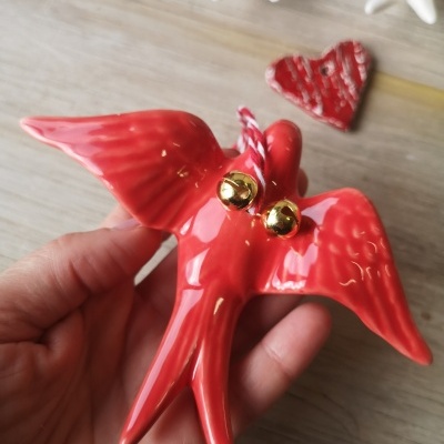 Ornamento vermelho em forma de pardal com sinos dourados, segurado por uma mão sobre mesa de madeira com coração vermelho ao fundo