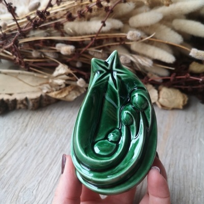 Escultura cerâmica verde de presépio com estrela, segurada por mão, ramos secos ao fundo